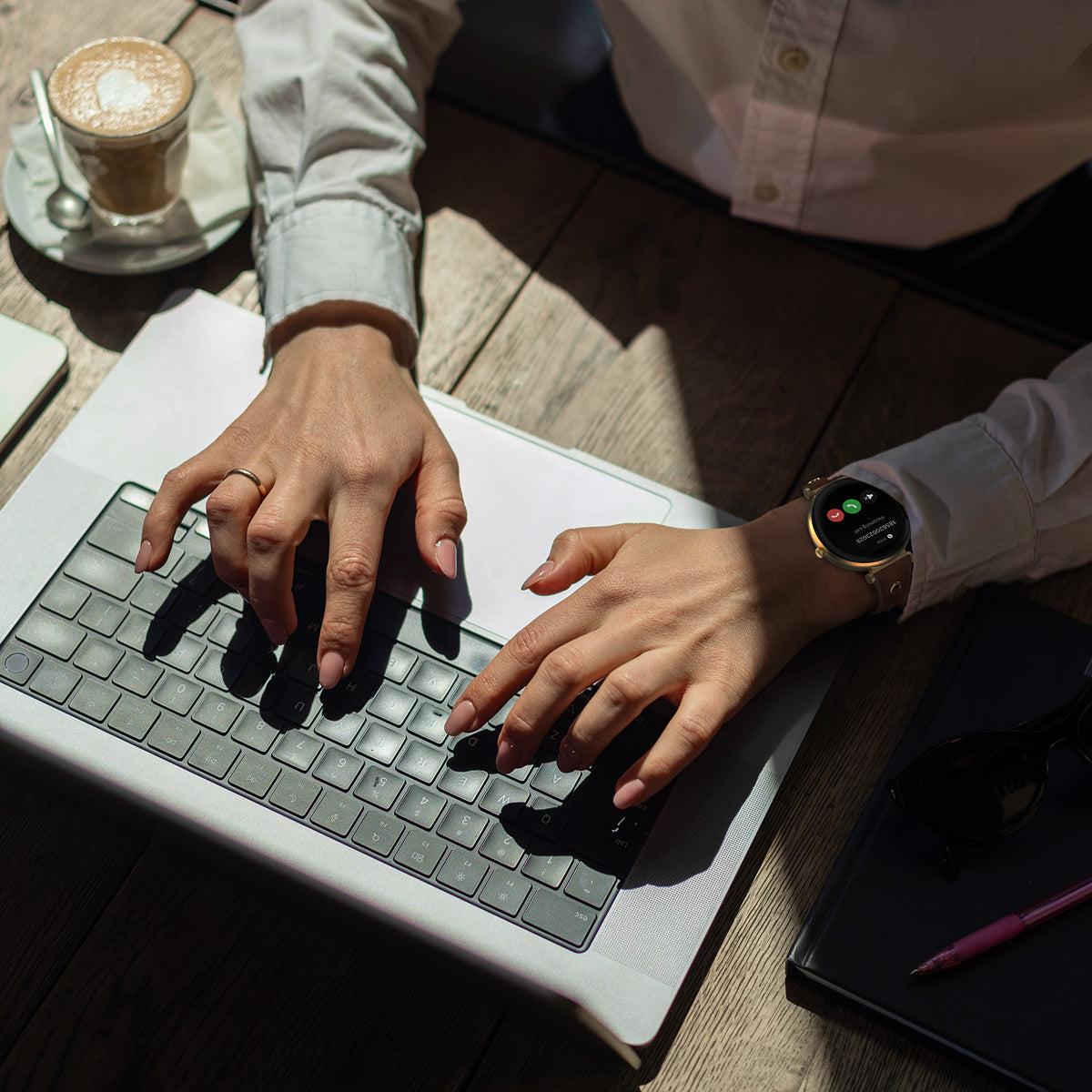 Person typing on a laptop with a cup of coffee and notebook in the background wearing smartwatch