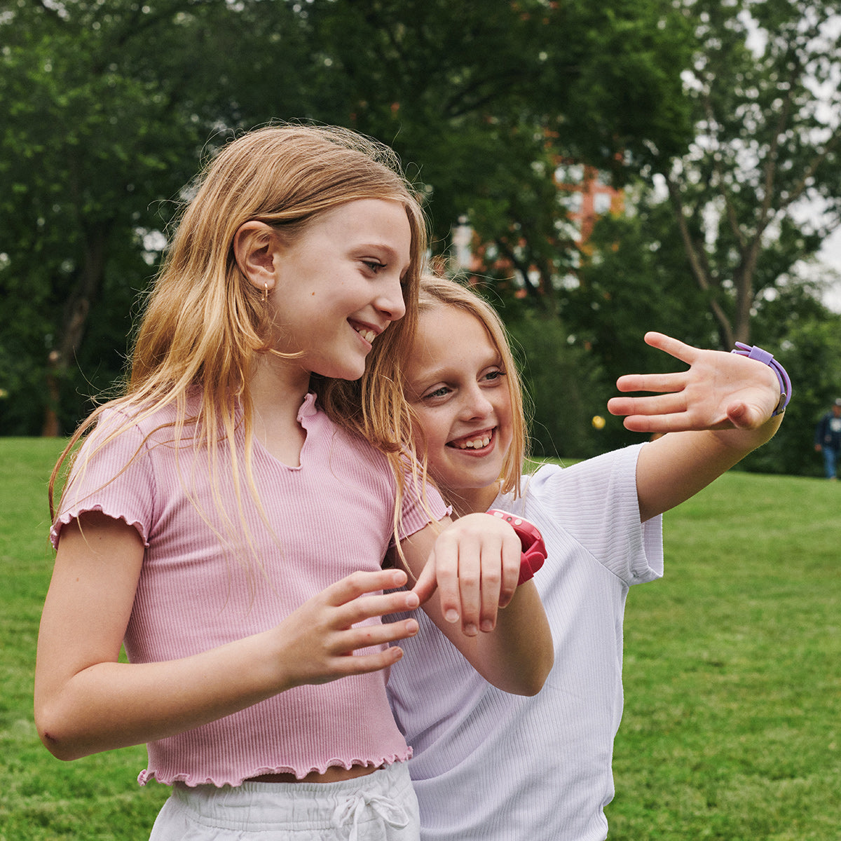 Two young girls standing outdoors in a park, smiling and interacting with each other looking at smartwatch