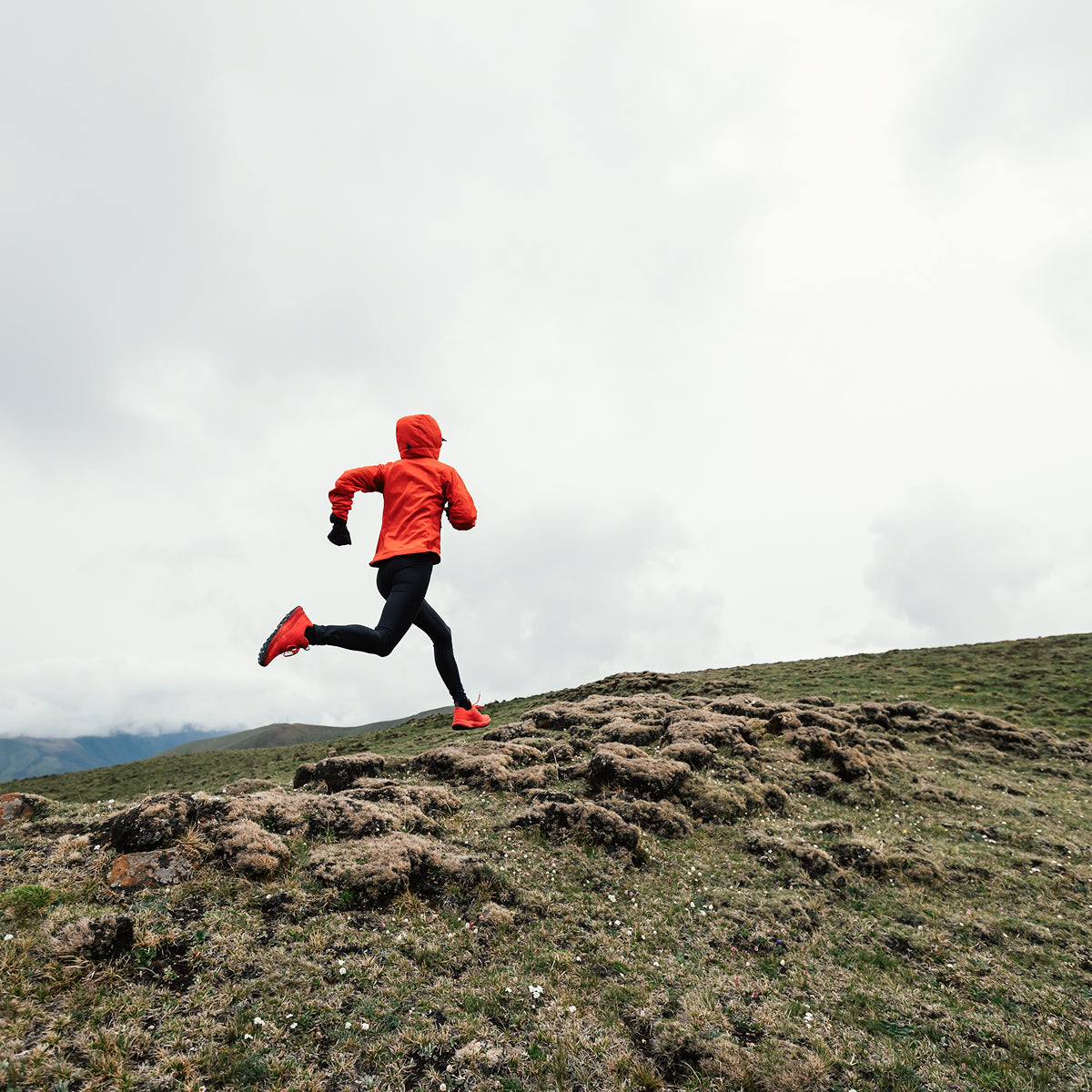Person running on a grassy hill with a cloudy sky wearing a red jacket