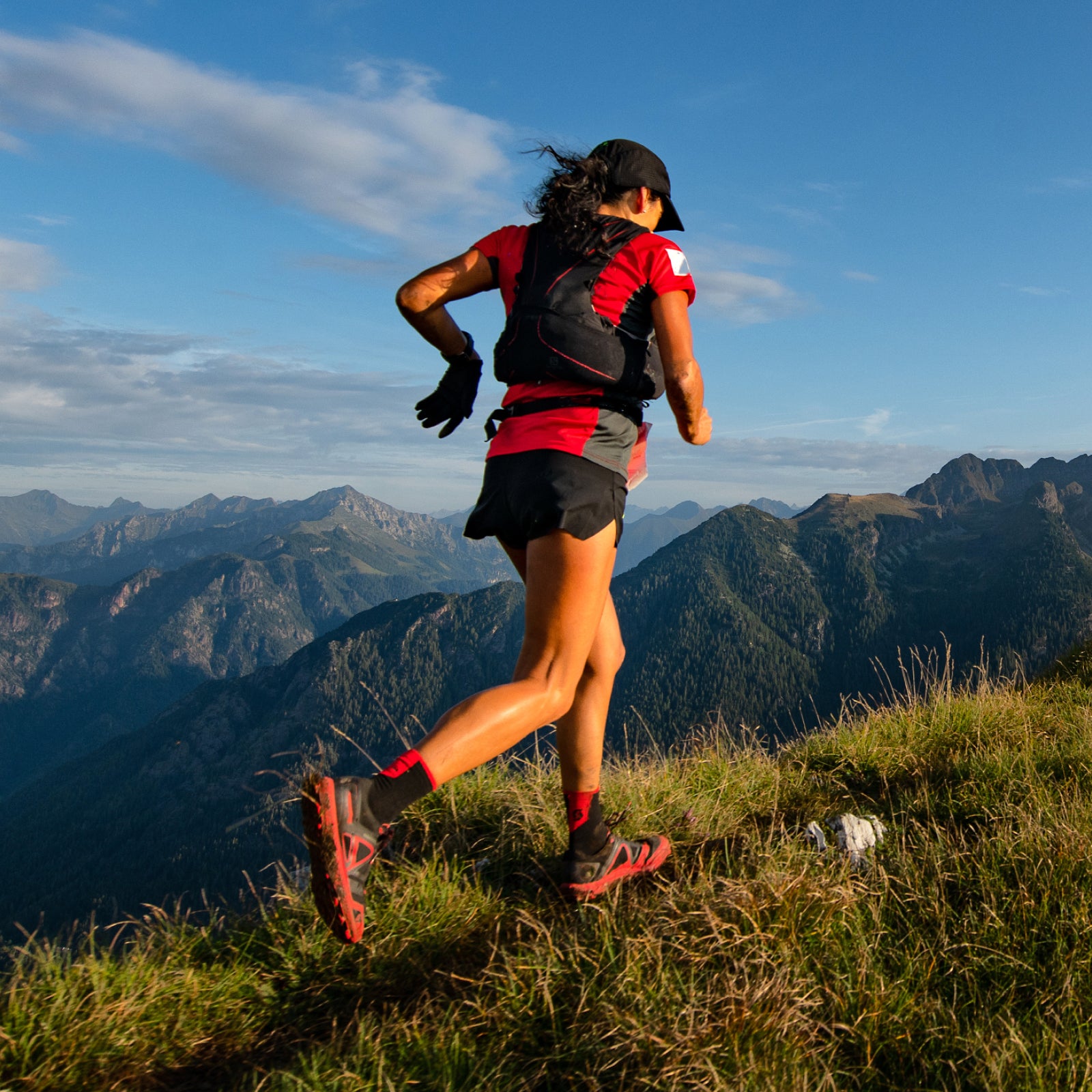 Person running on a mountain trail with a backpack, surrounded by mountains and blue sky.